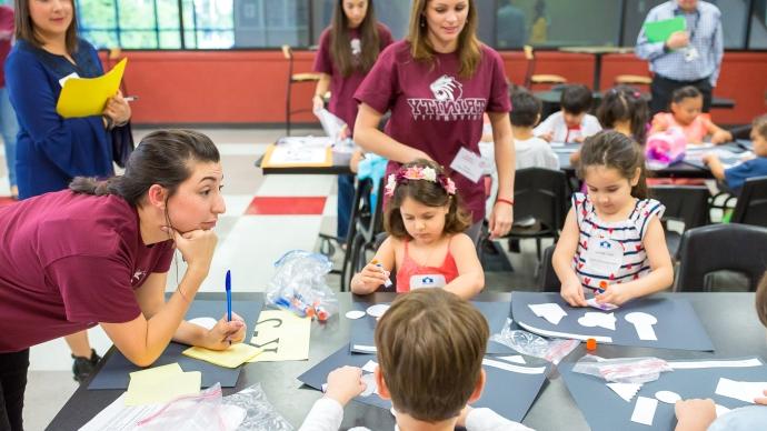 Two Trinity MAT intern teachers stand with a table of K-2 students working on a paper craft