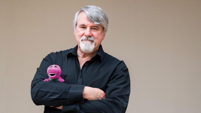 bob west in black shirt standing in front of a tan background with his arms crossed, holding a barney plush doll