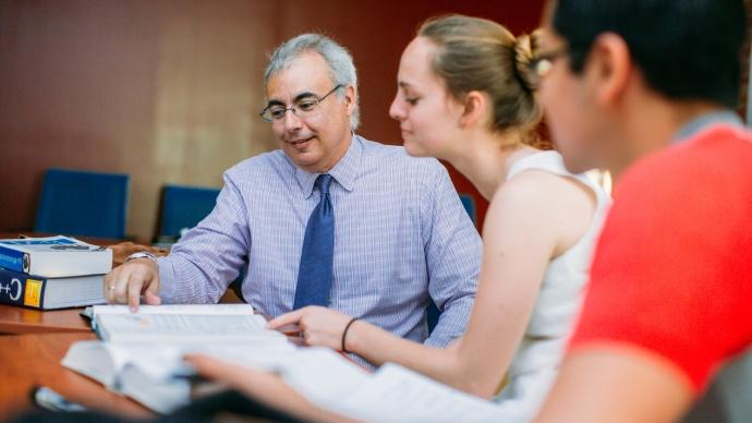 Professor sitting with two students, teaching from books 